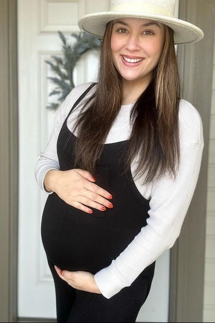 woman wearing a black maternity overall dress and a white long sleeve top, showcasing her baby bump while smiling