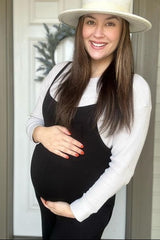 woman wearing a black maternity overall dress and a white long sleeve top, showcasing her baby bump while smiling