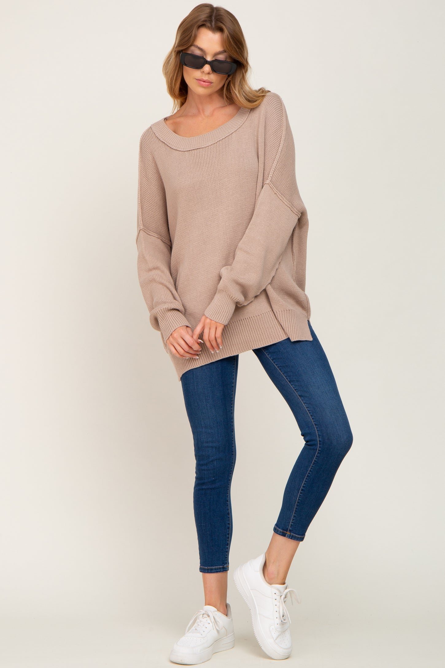 woman wearing a beige oversized sweater with long sleeves and blue skinny jeans, paired with white sneakers and sunglasses