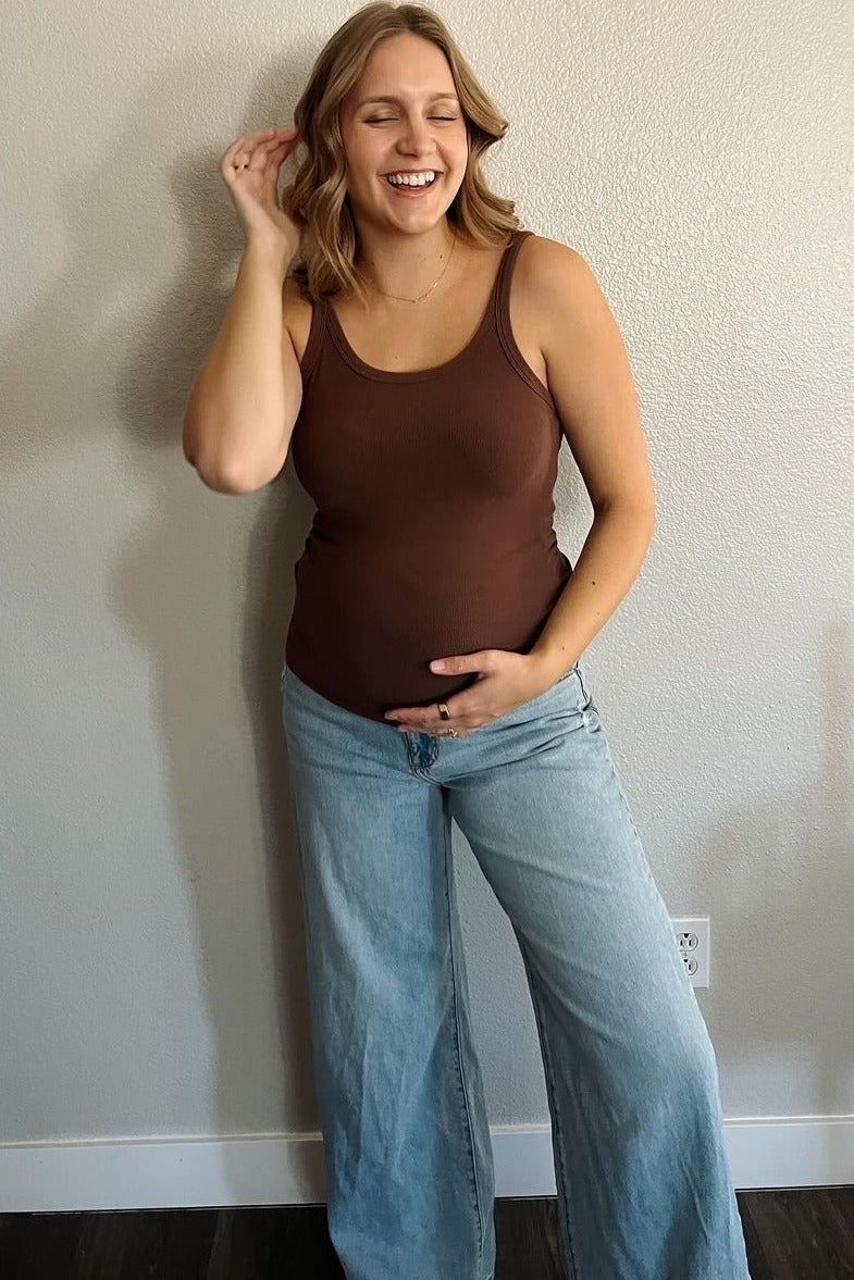 Smiling pregnant woman in brown tank top and wide leg jeans holding her baby bump against a neutral wall