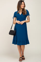 woman wearing a teal maternity dress with short sleeves, cinched waist, and ruffle hem, holding a black handbag