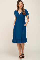 woman wearing a teal maternity dress with puff sleeves, a ruffled hem, and pockets, showing her baby bump