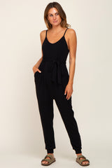woman wearing a black sleeveless jumpsuit with a tie waist and pockets, styled with brown sandals