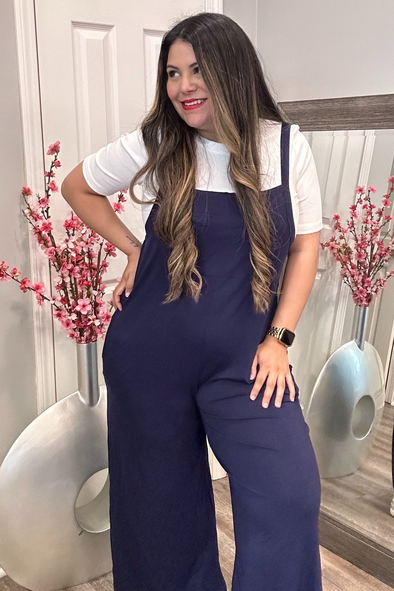 woman wearing a navy blue wide-leg jumpsuit over a white shirt, posing in a stylish interior with floral decor