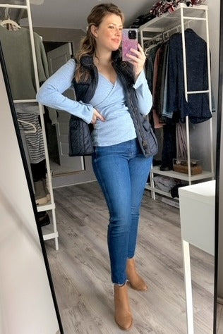 woman in blue wrap top, navy quilted vest, skinny jeans, and tan ankle boots taking a mirror selfie in a closet