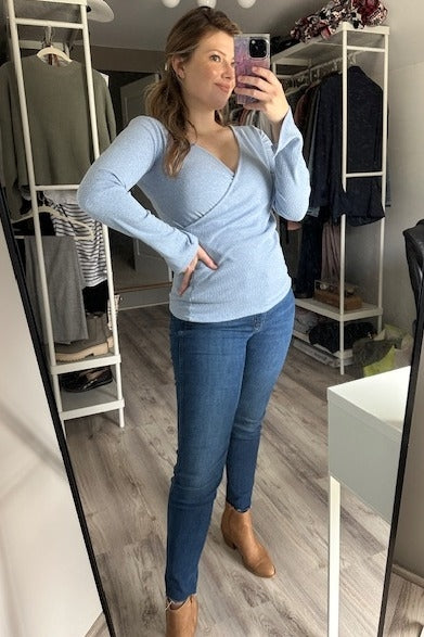 woman wearing a light blue long sleeve wrap top with blue jeans and tan ankle boots, posing in front of a mirror