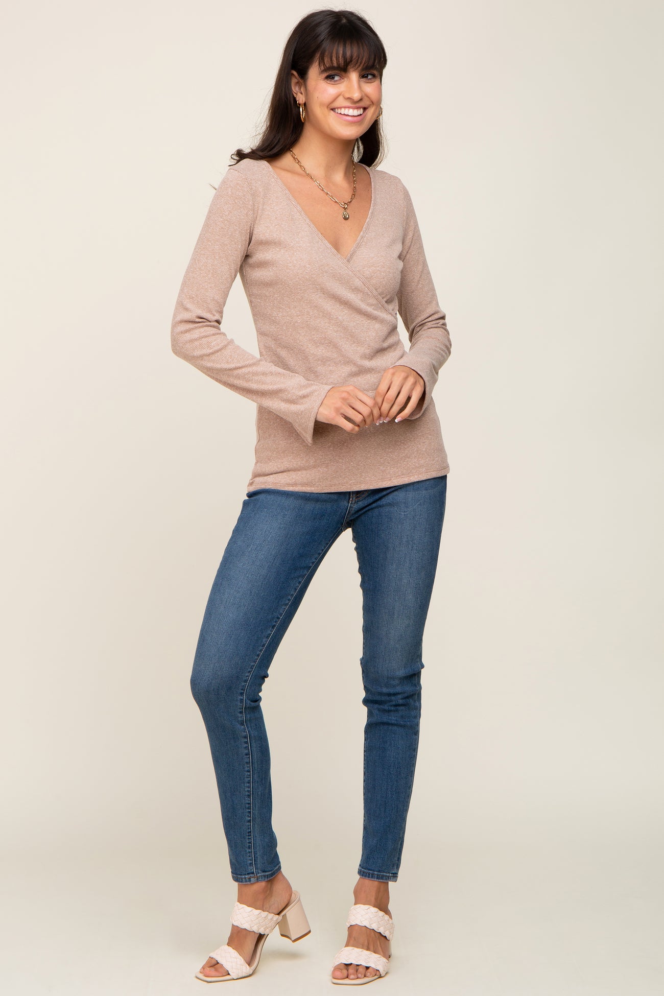 woman wearing a beige long sleeve wrap top paired with blue skinny jeans and white heeled sandals