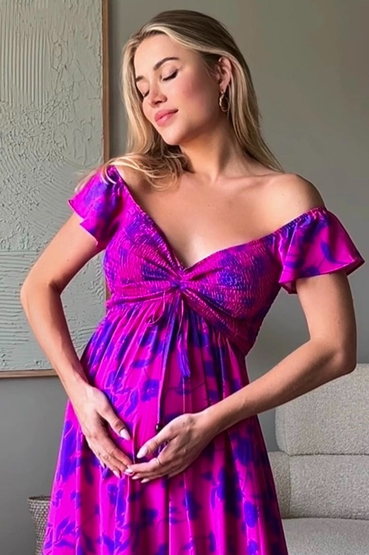 woman wearing a vibrant pink off-shoulder maternity dress with ruffle sleeves, holding her baby bump