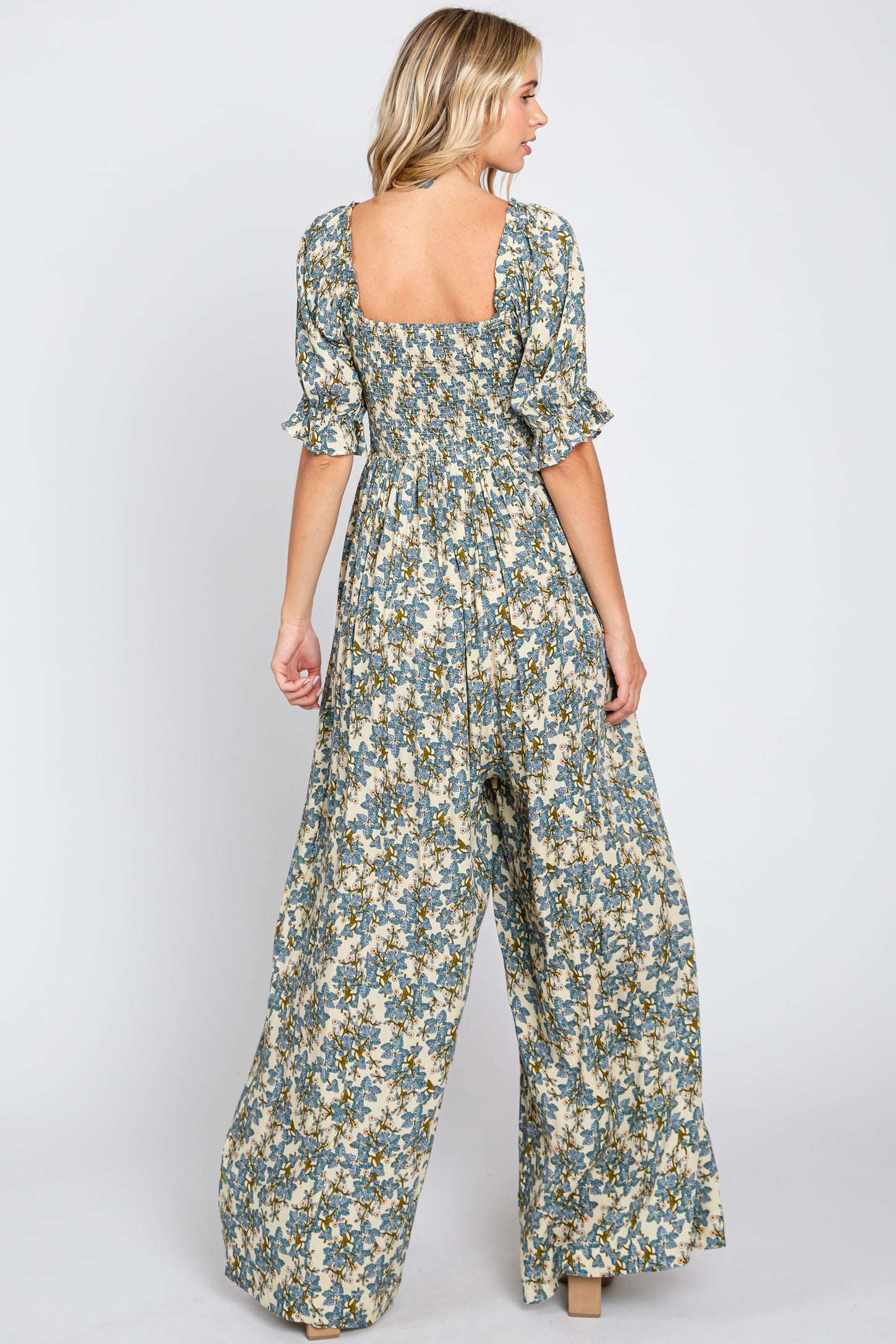Back view of a woman wearing a blue floral print wide leg jumpsuit with smocked bodice and ruffled sleeves
