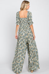 Back view of a woman wearing a blue floral print wide leg jumpsuit with smocked bodice and ruffled sleeves
