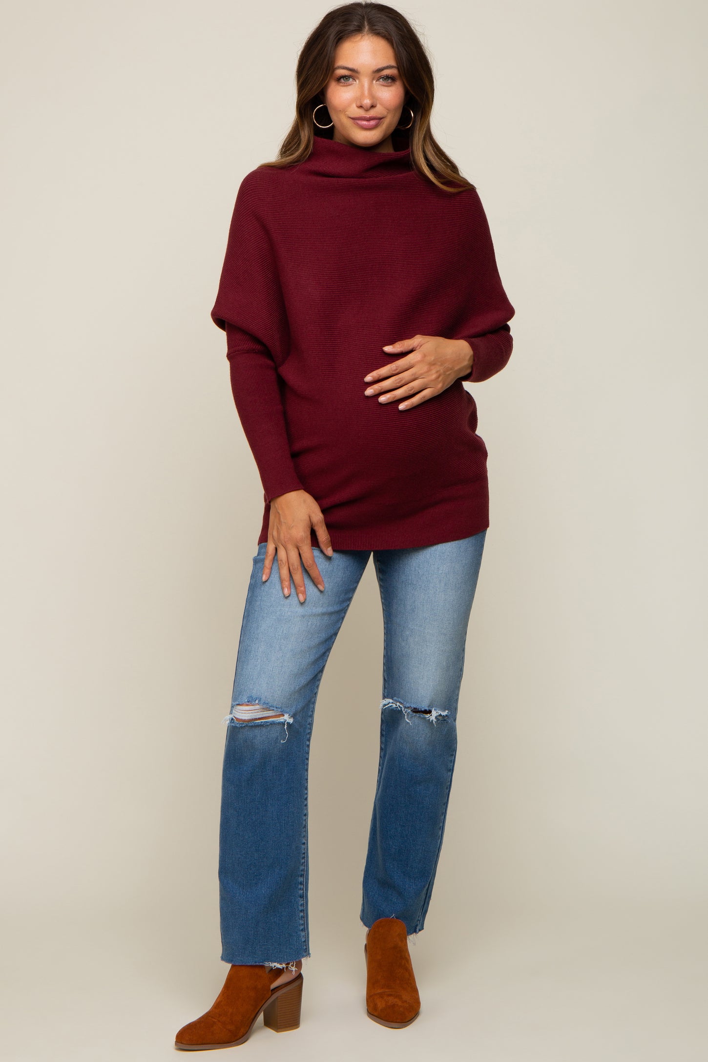 woman wearing a burgundy oversized maternity sweater with a cowl neck and holding her baby bump, paired with distressed jeans and brown ankle boots