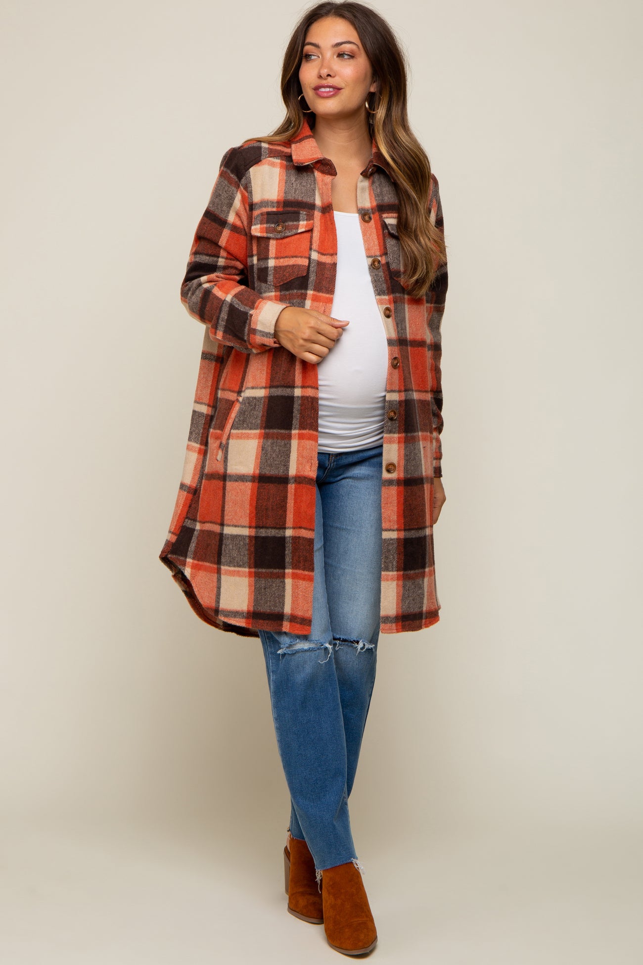 woman wearing a plaid maternity shirt jacket in orange, black, and cream, paired with distressed jeans and brown ankle boots, showing baby bump