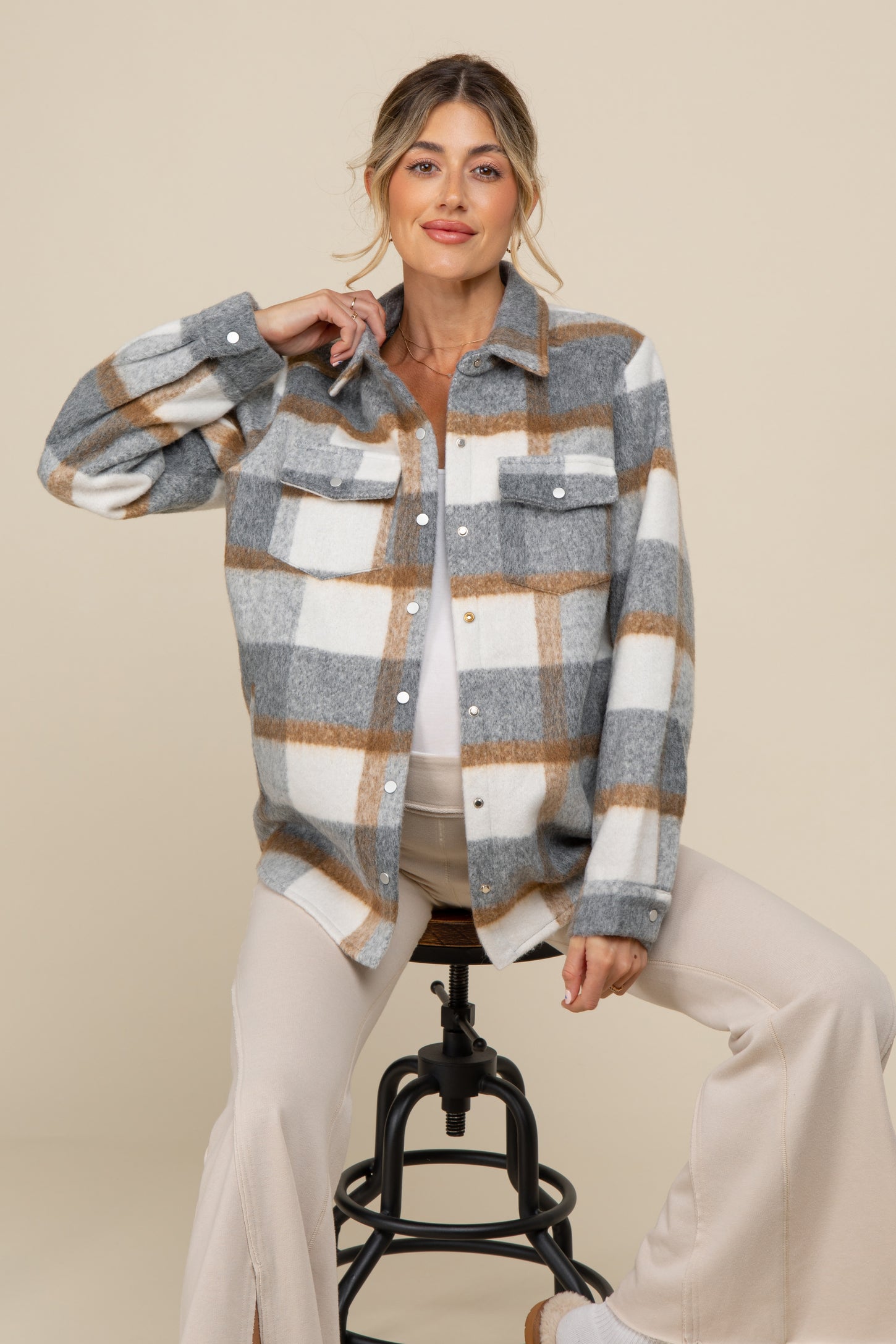 woman wearing a gray and white plaid maternity shirt jacket over a light top, sitting on a stool with a relaxed pose