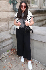 woman wearing a striped short sleeve top, black wide-leg pants, and white sneakers, with a striped sweater draped over her shoulders
