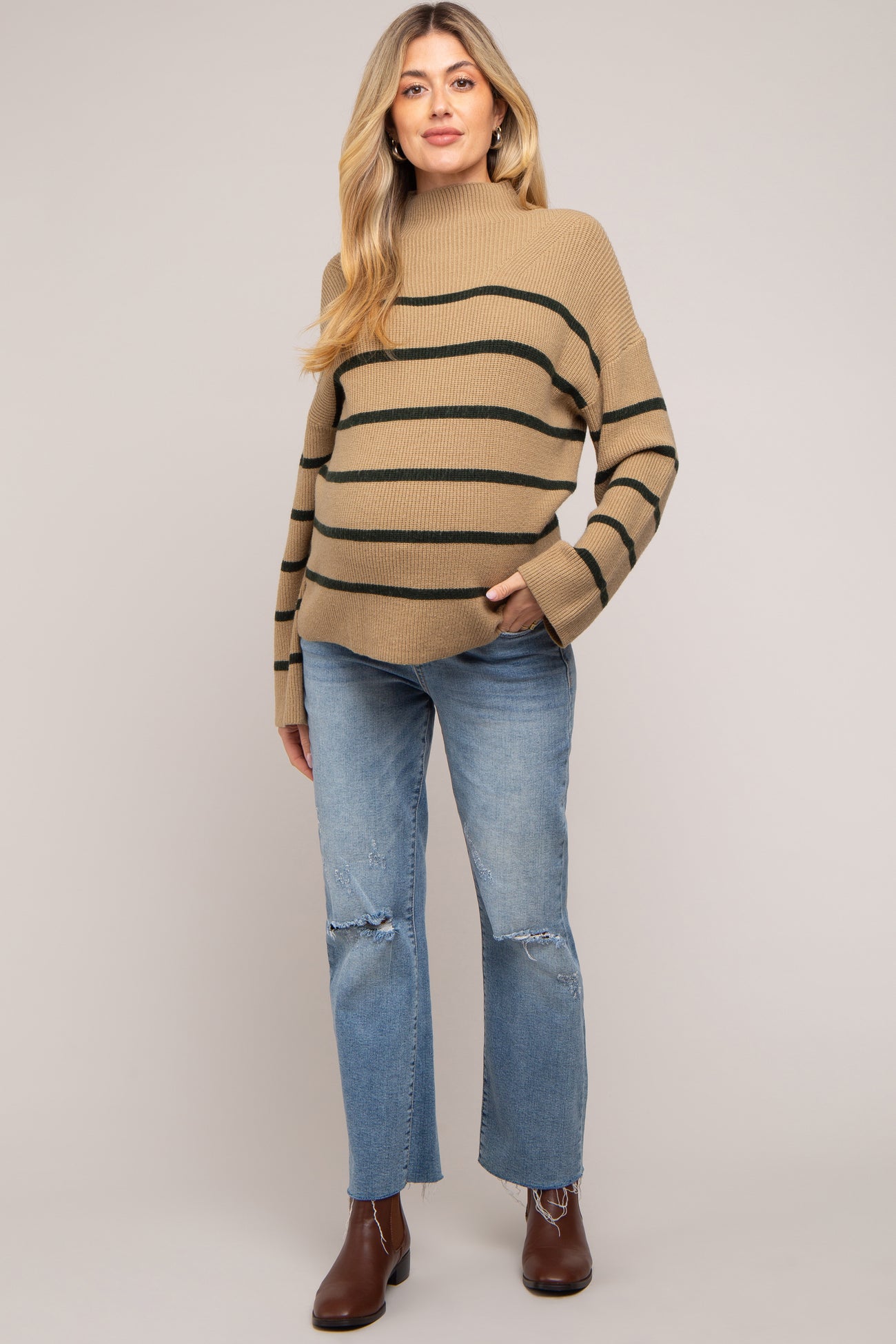 woman wearing a tan striped maternity sweater and distressed jeans, holding her baby bump