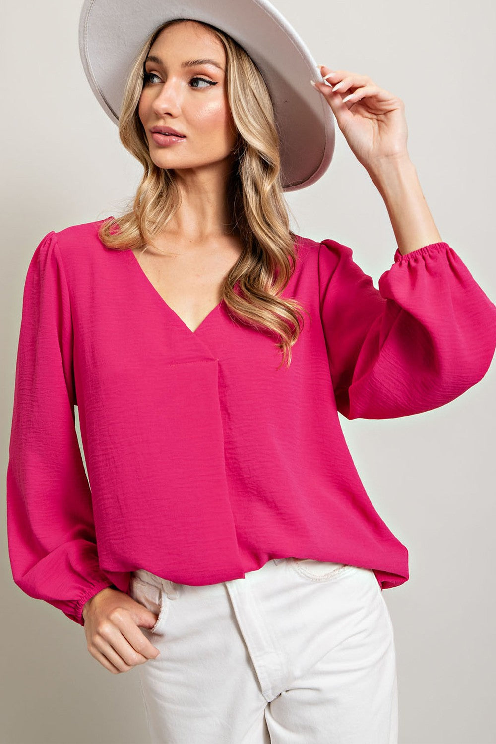 woman wearing a bright pink v-neck blouse with balloon sleeves and white pants, accessorized with a wide-brimmed hat