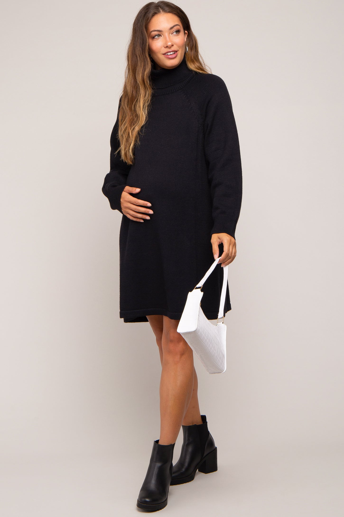 woman wearing a black knit maternity sweater dress and holding her baby bump with a white handbag