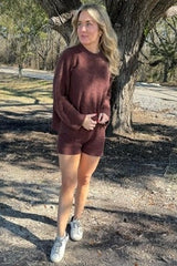 woman wearing a maroon sweater and matching shorts, standing outdoors near a tree, with sneakers on