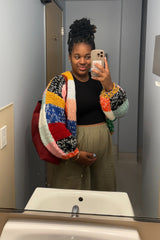 woman wearing a black maternity top and an oversized multicolored cardigan, holding her baby bump in a mirror selfie