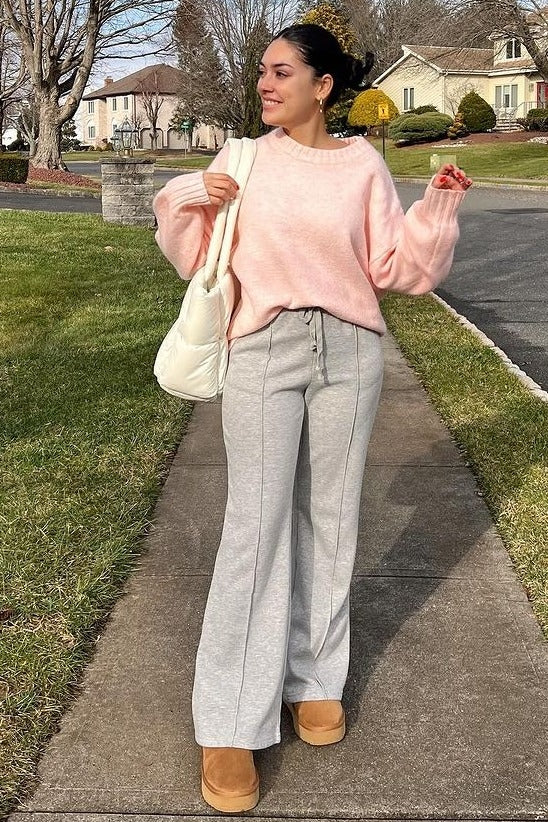woman wearing a light pink oversized sweater, gray wide-leg sweatpants, and tan boots, holding a cream shoulder bag