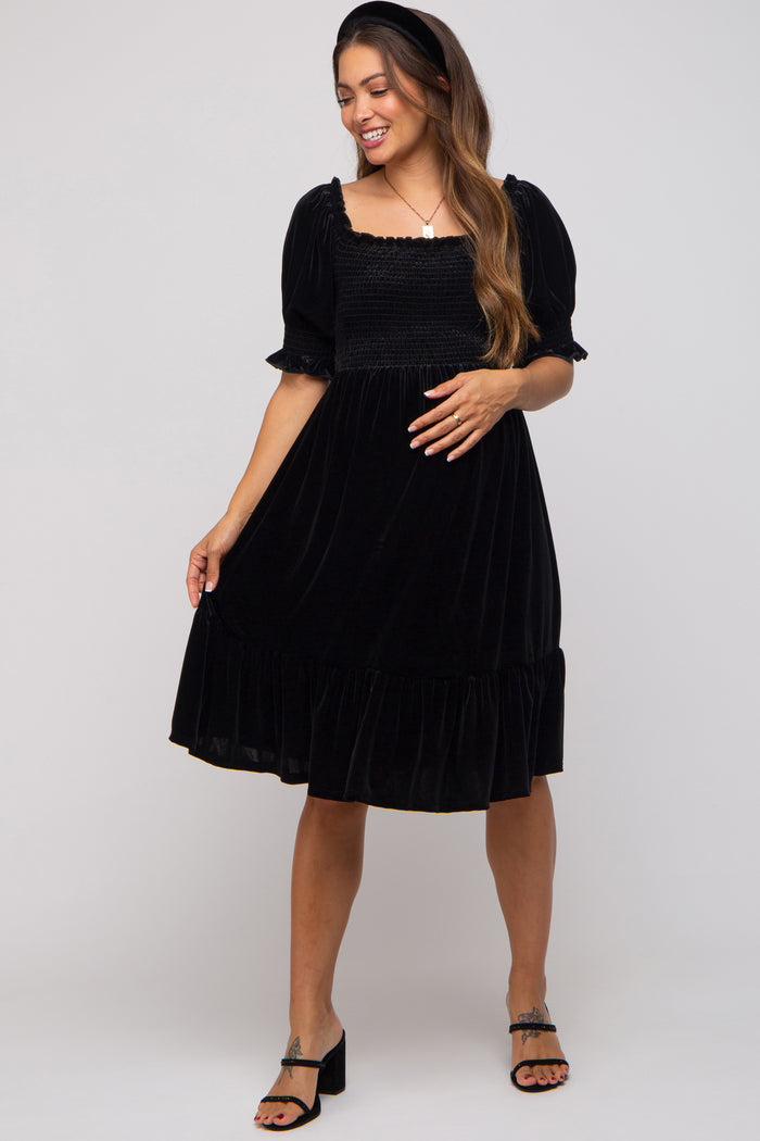 woman wearing a black velvet maternity dress with puff sleeves and a ruffled hem, posing with one hand on her hip