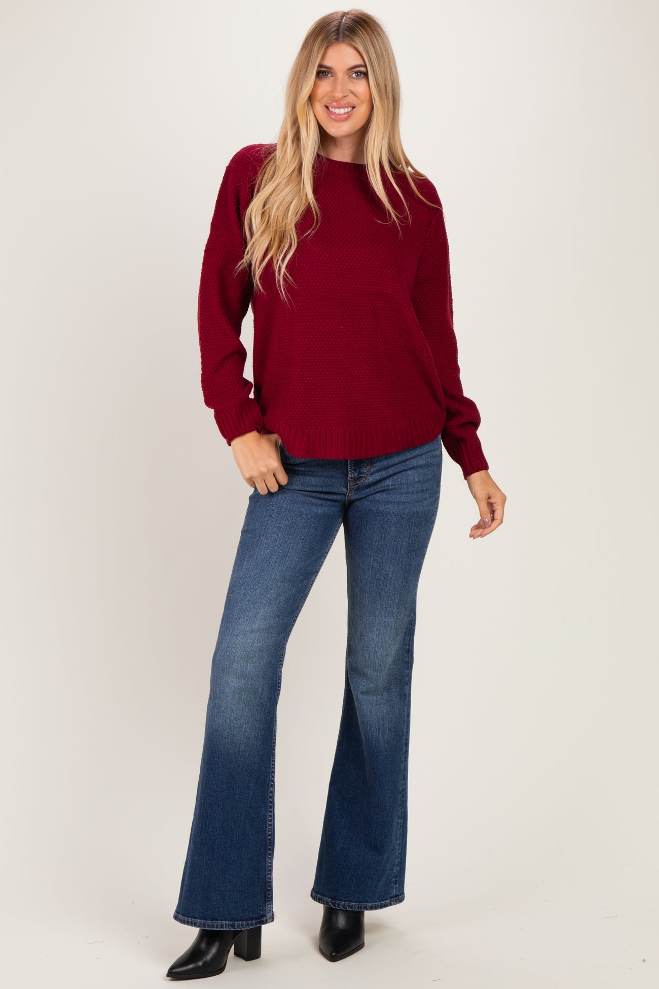 woman wearing a burgundy oversized knit sweater and flared jeans, standing with one hand on her hip