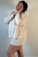 woman wearing a beige knit sweater and matching mini skirt, smiling and posing with hands near her face