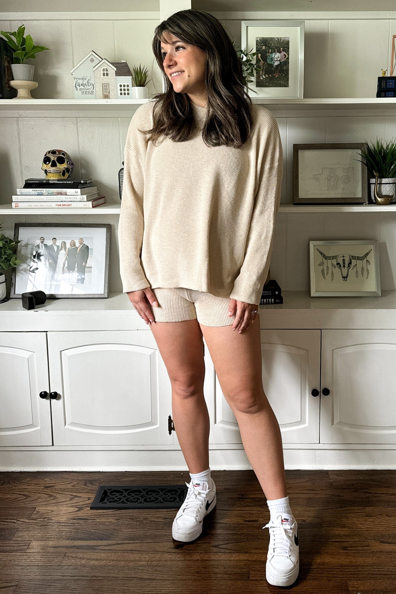 woman wearing a cream oversized sweater and matching knit shorts, styled with white sneakers and standing in a well-decorated room