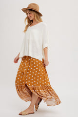 woman wearing a white oversized knit top and a brown floral maxi skirt, accessorized with a tan hat and sandals
