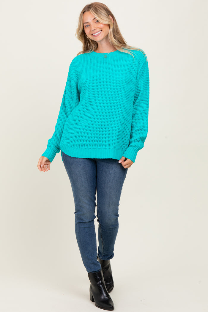 woman wearing a bright turquoise oversized knit sweater and blue skinny jeans, standing with one hand on her hip