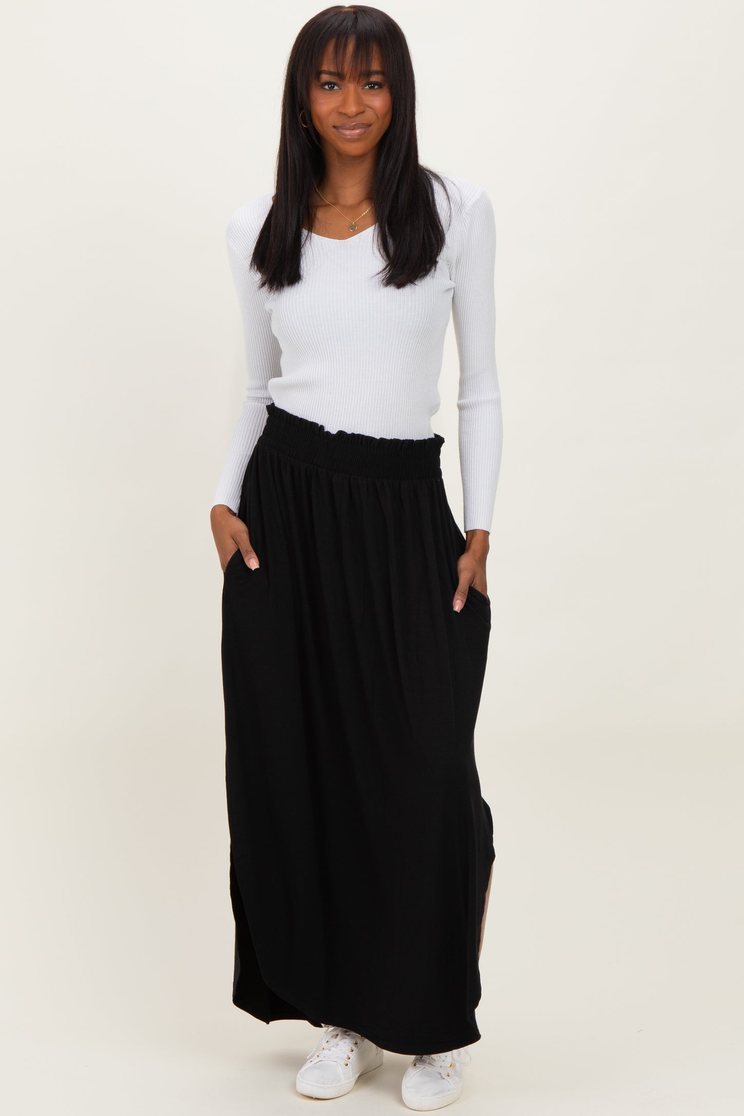 woman wearing a white ribbed long sleeve top and a black maxi skirt with pockets, standing against a neutral background