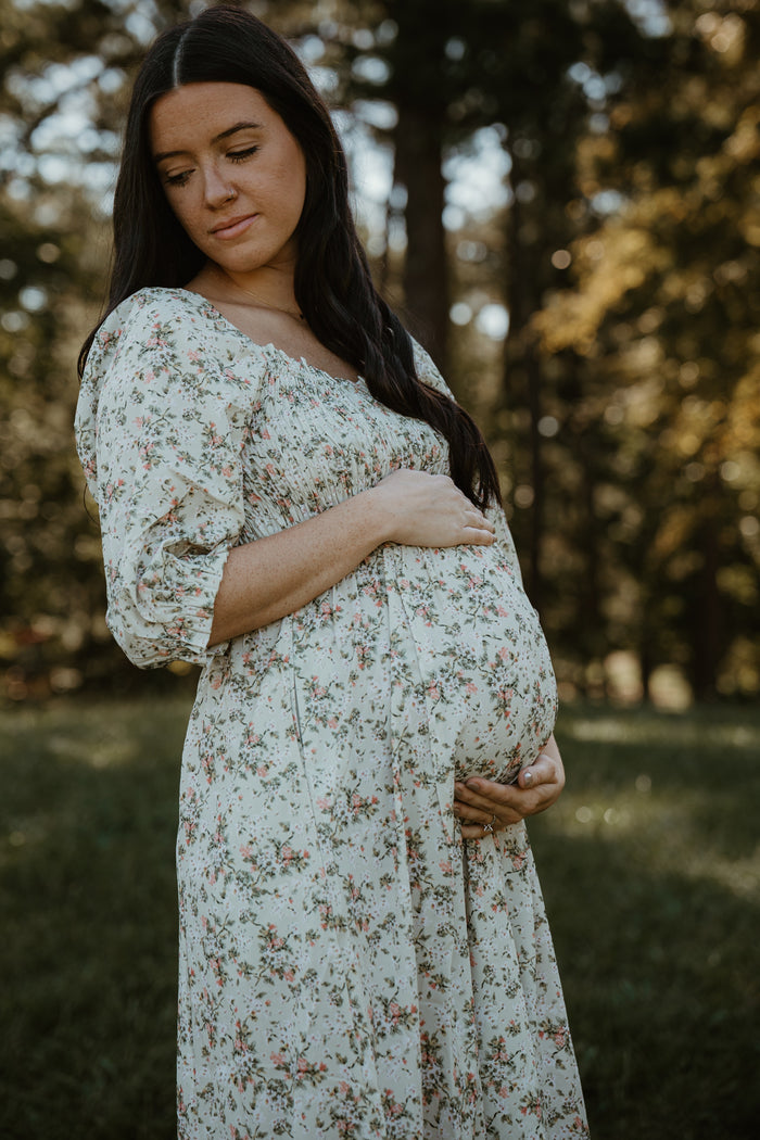 woman wearing a floral maternity dress and gently cradling her baby bump in a sunny outdoor setting