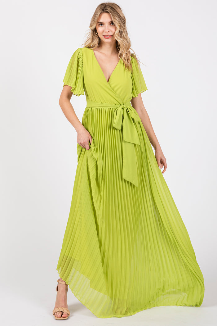 woman wearing a pleated bright green maxi dress with a v-neck and tie belt, standing with hands in pockets