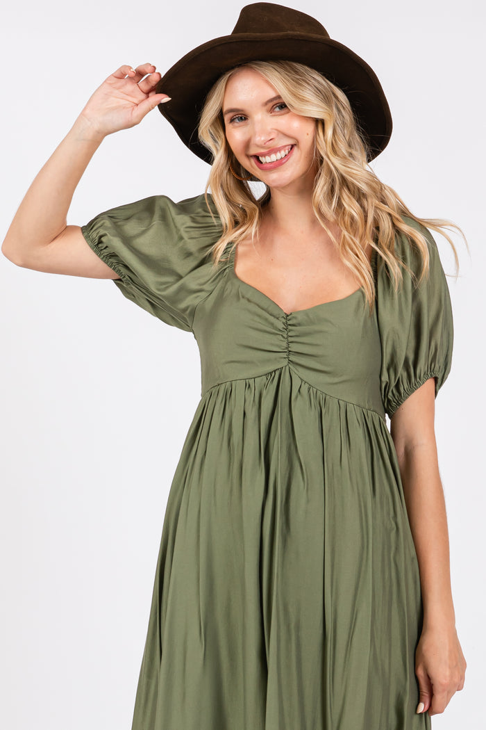 woman wearing an olive green puff sleeve maxi dress and a brown hat, smiling and posing with one hand raised