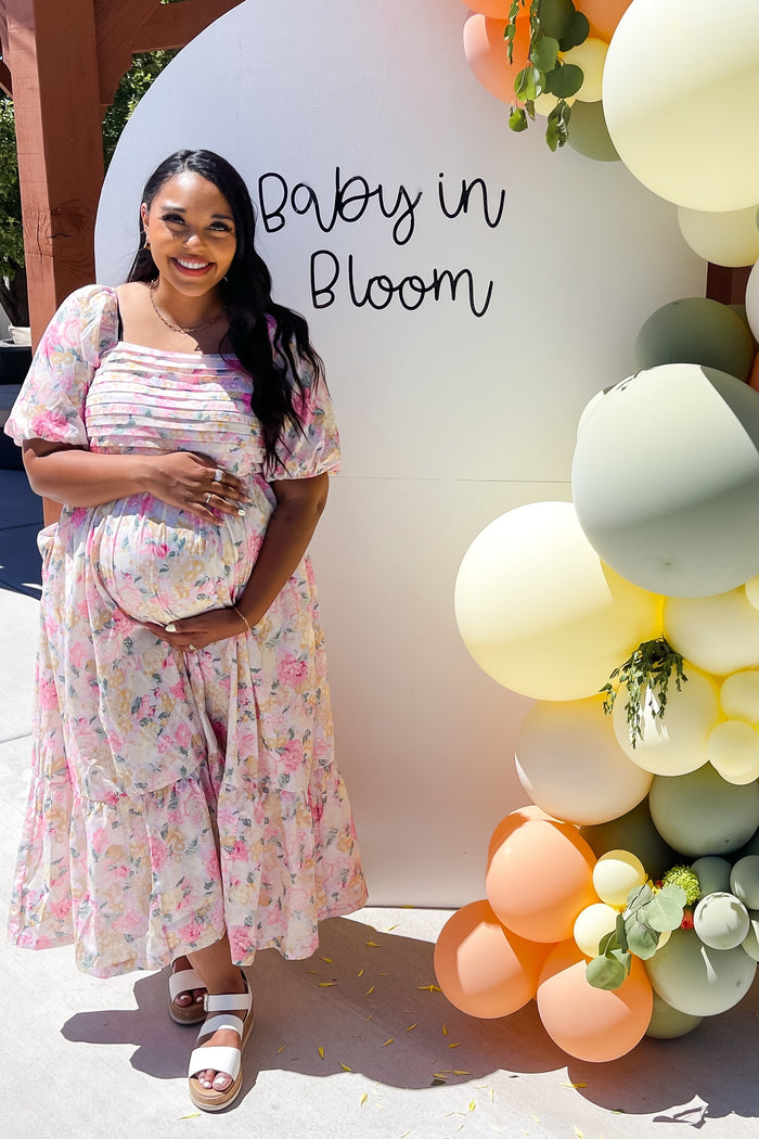 woman wearing a floral maternity dress with a baby bump, standing next to a balloon display that says "Baby in Bloom