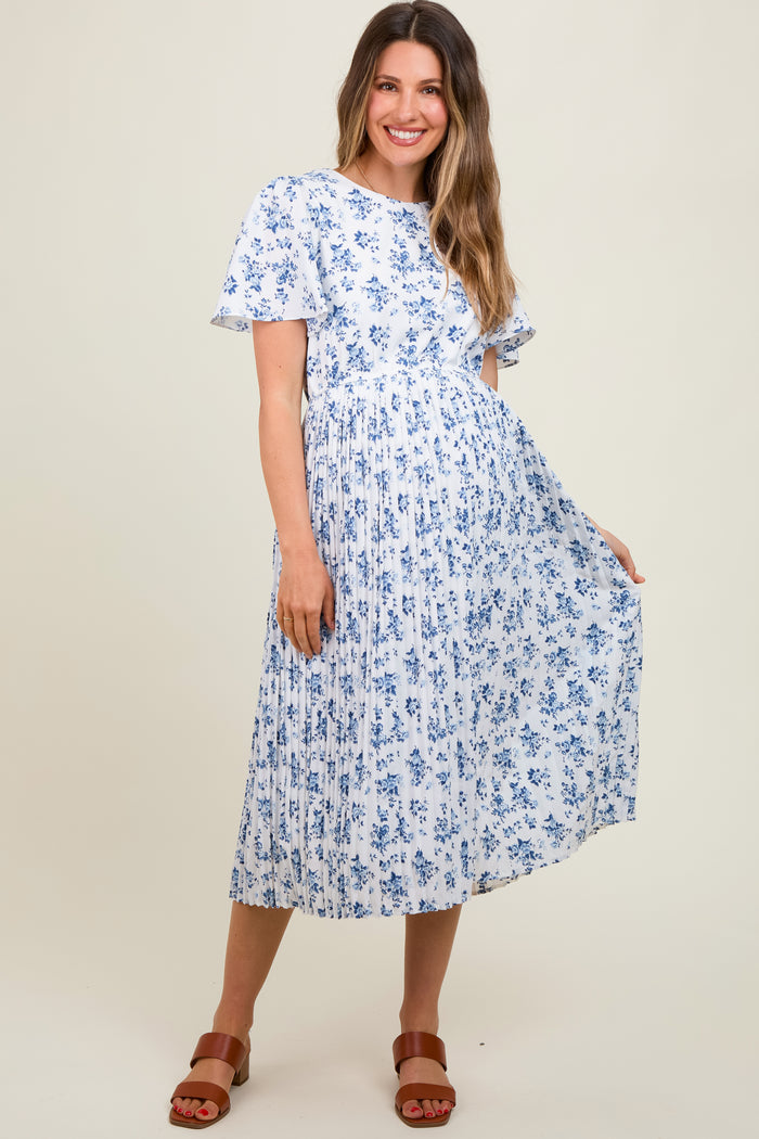 woman wearing a white floral maternity dress with pleats and flutter sleeves, standing and smiling