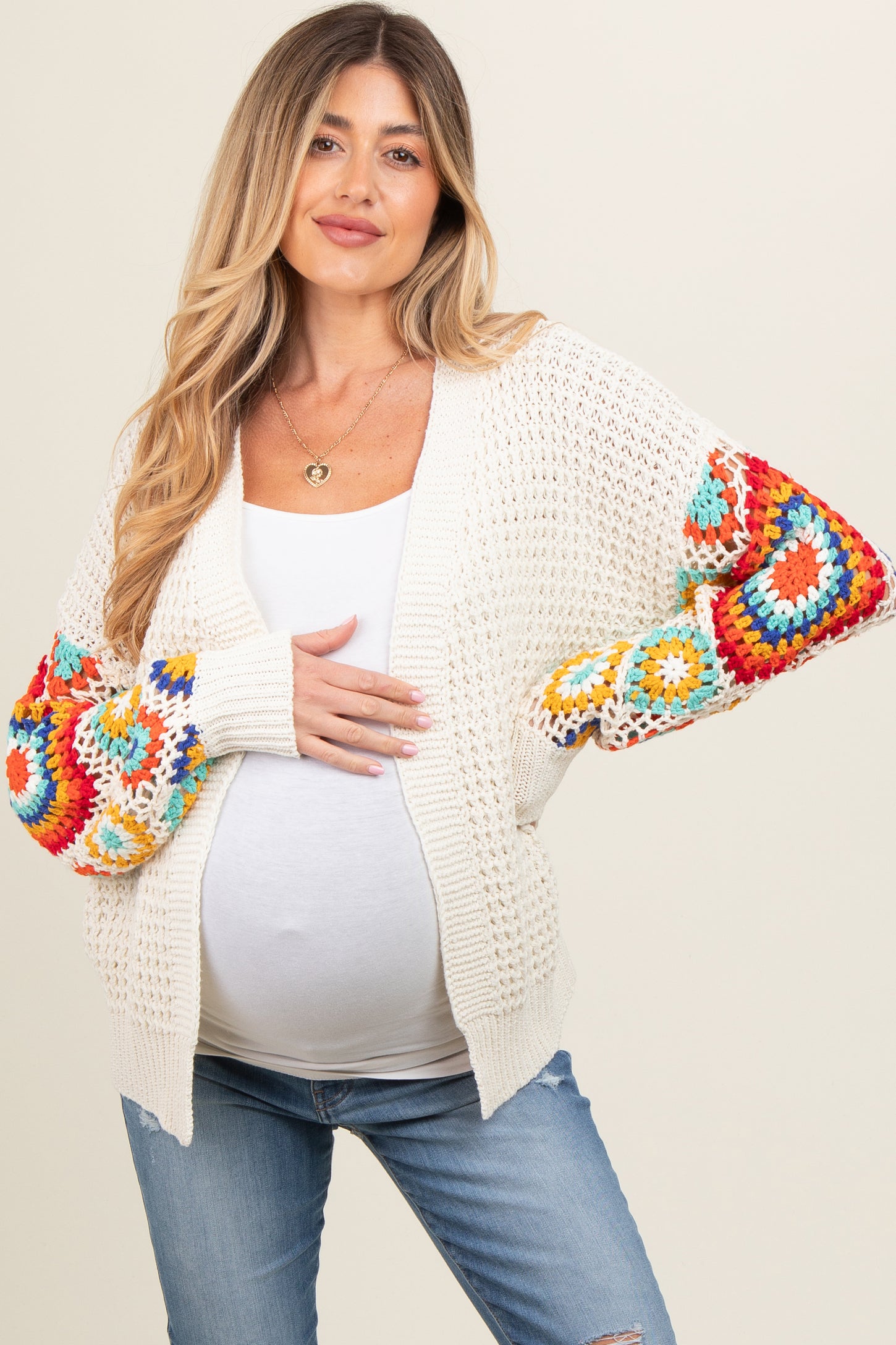 woman wearing a cream crochet maternity cardigan with colorful patterned sleeves and holding her baby bump