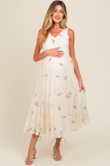 woman wearing a cream maternity dress with floral embroidery and tiered ruffles, holding her baby bump