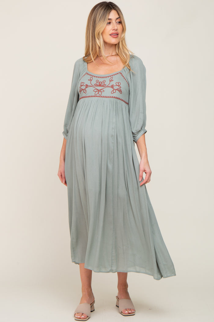 pregnant woman wearing a sage green maternity midi dress with floral embroidery and three-quarter sleeves